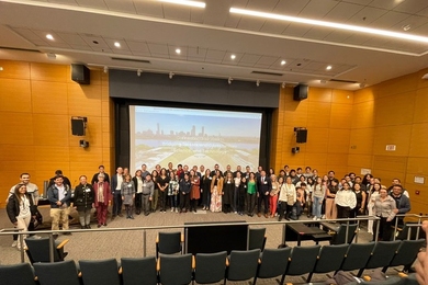 About 65 people pose standing in a semicircle on a large auditorium stage, with the backs of empty seats in the foreground and a video screen behind them.